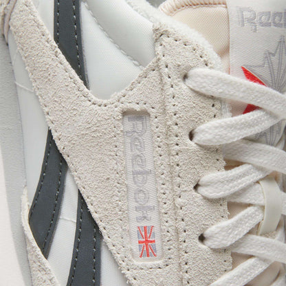 Close-up of the Women's Classic AZ Shoes by Reebok in white and beige, featuring suede and leather accents plus a woven logo tag with a small Union Jack near the laces—ideal for vintage sneaker enthusiasts.