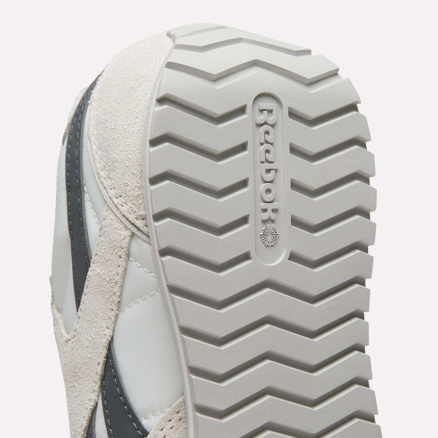 Close-up of the sole on Women's Classic AZ Shoes by Reebok, highlighting the zigzag tread and embossed Reebok logo. These vintage sneakers come in light gray and beige.