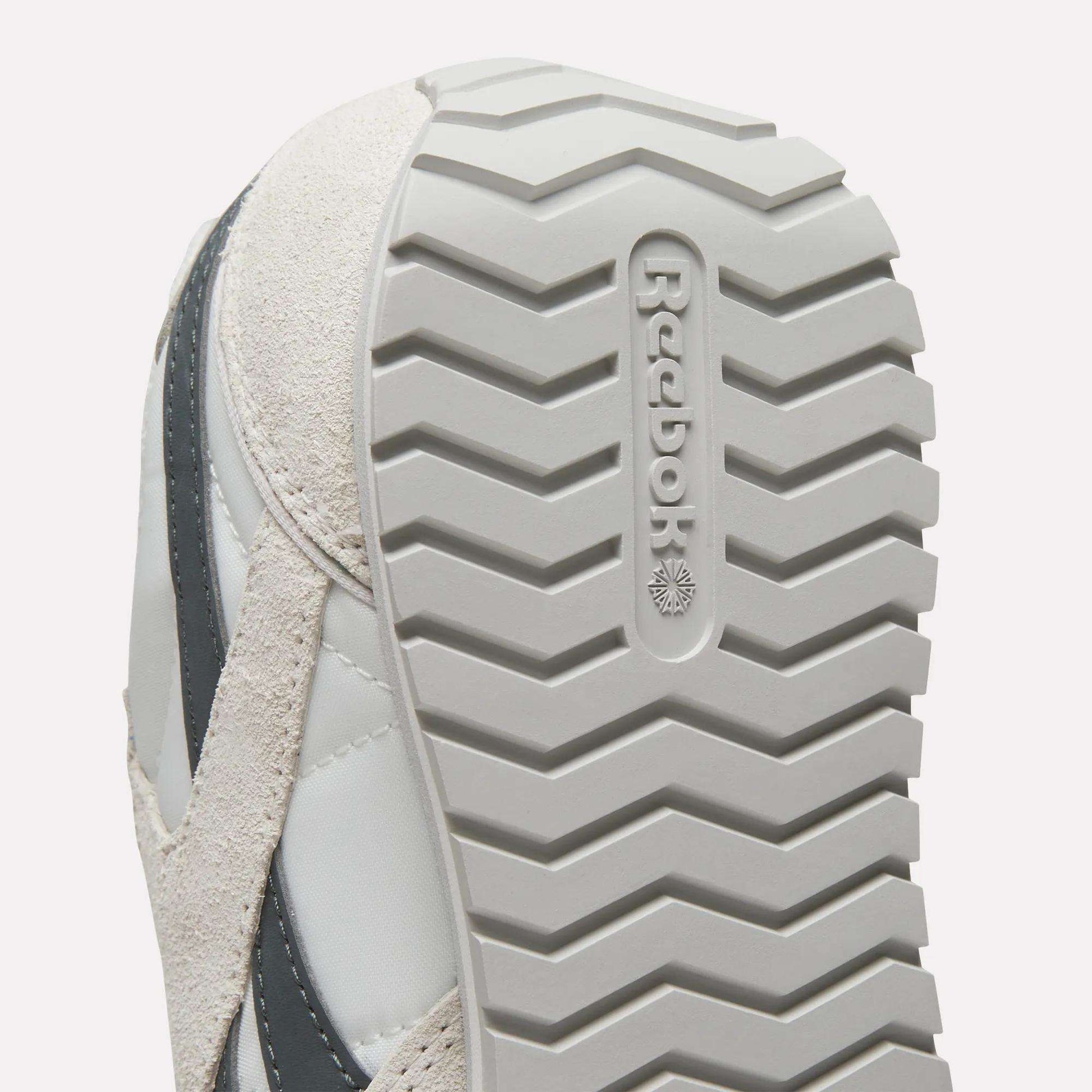 Close-up of the sole on Women's Classic AZ Shoes by Reebok, highlighting the zigzag tread and embossed Reebok logo. These vintage sneakers come in light gray and beige.