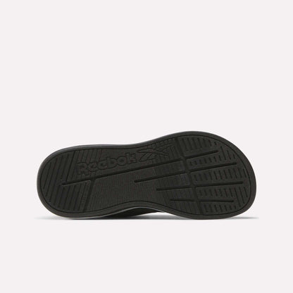 The image shows the black rubber outsole of the Reebok Nano Play Stretch Lace And Top Strap Shoes for little kids, with textured grip patterns and an embossed Reebok logo featuring the brand's vector symbol in the center.