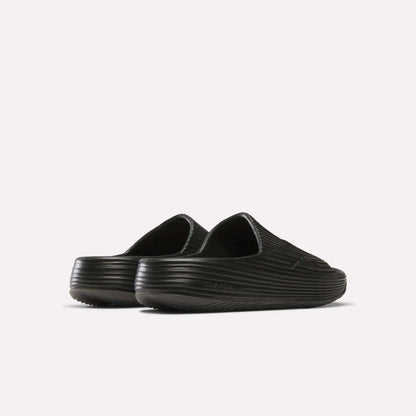 The Reebok DMX Recovery Walking Slides are black slip-on clogs with a ribbed texture, closed toes, and super-soft foam for extra comfort, shown from the back and side on a plain white background.