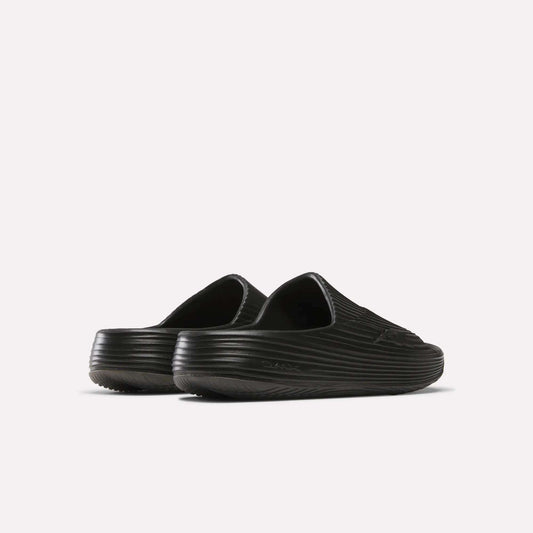 The Reebok DMX Recovery Walking Slides are black slip-on clogs with a ribbed texture, closed toes, and super-soft foam for extra comfort, shown from the back and side on a plain white background.