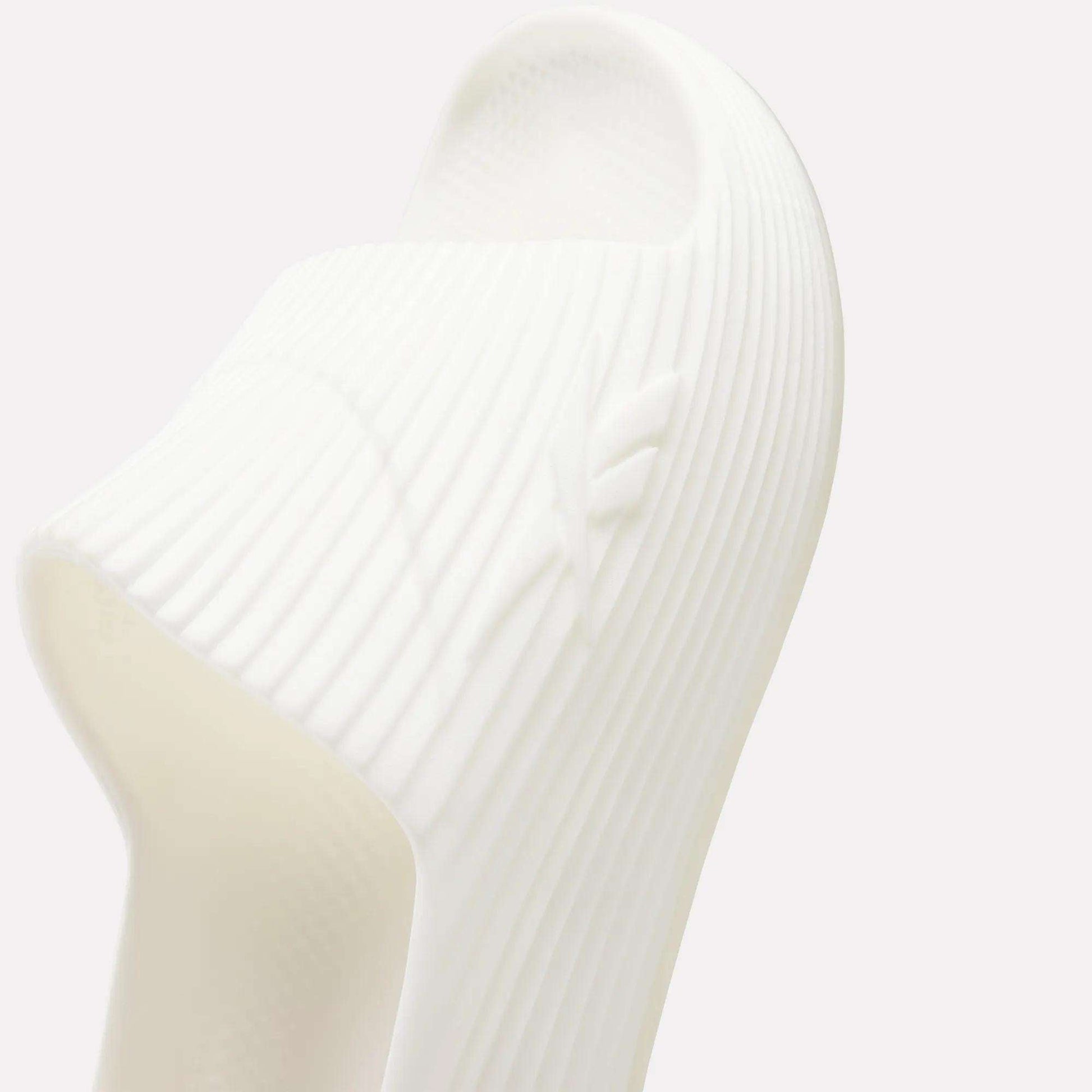 Close-up of white Reebok DMX Recovery Walking Slides featuring ribbed design, subtle embossed logo on the side, and air cushioning, photographed against a light background.