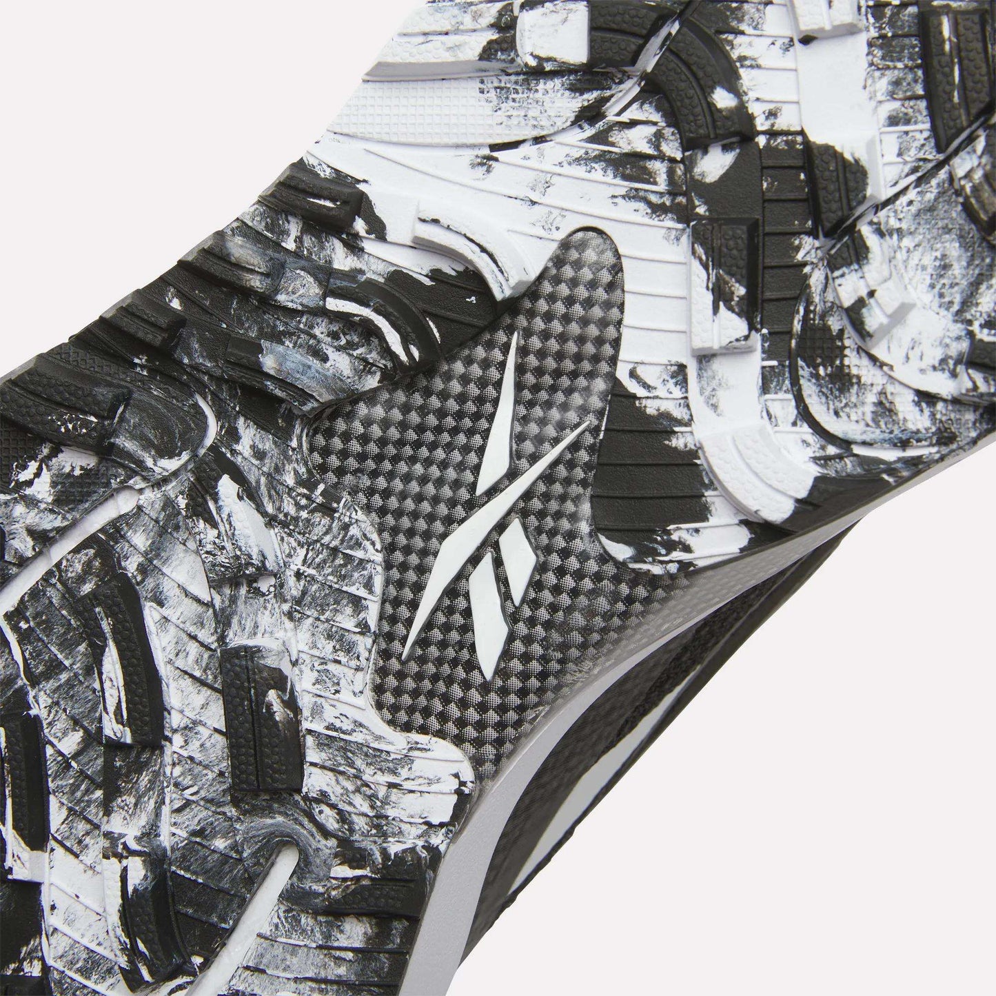 Close-up of the Reebok Nano Golf Shoes sole, showing a textured black and white pattern with a central Reebok logo. Rugged, angular tread shapes enhance traction, while performance foam offers comfort on the course.