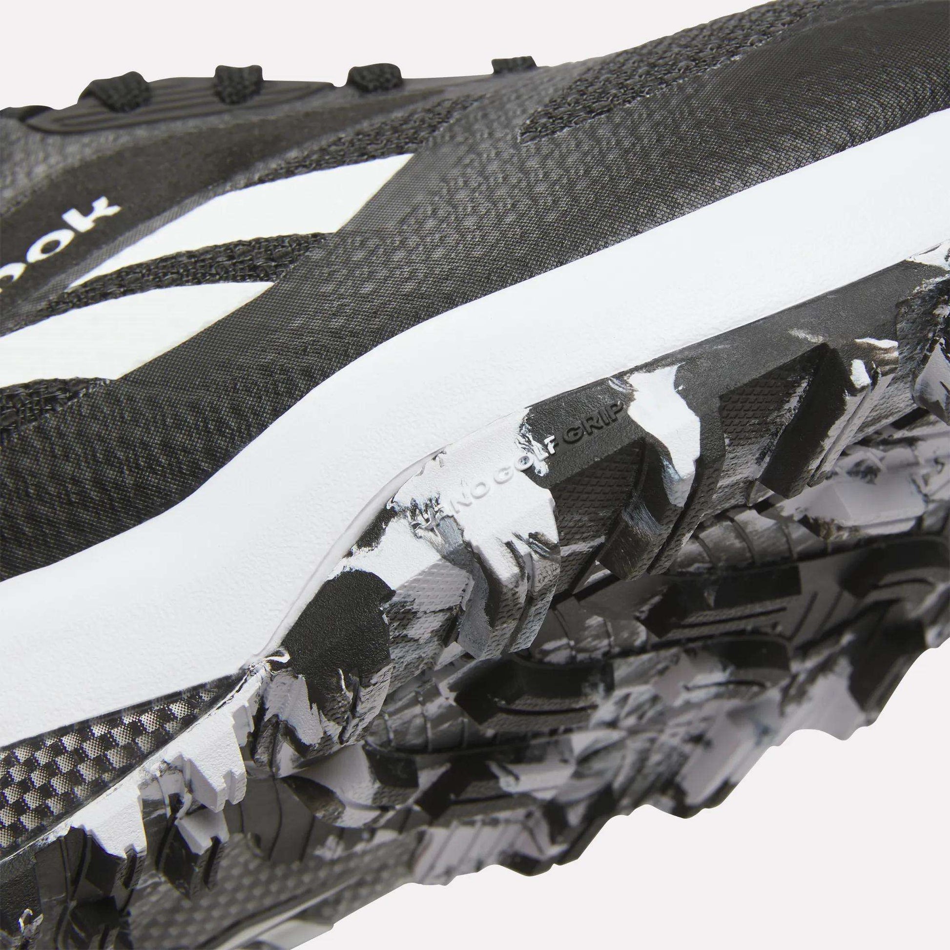 Close-up of Reebok Nano Golf Shoes in black and white, highlighting the textured sole with rugged tread pattern and FlexWeave upper for enhanced durability, plus a visible white logo.