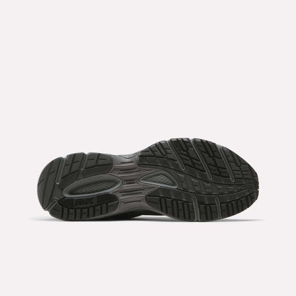 The image shows the black rubber outsole of Reebok Premier Trinity Shoes, viewed from the bottom. The tread has patterns and grooves for traction and flexibility, resembling retro running shoes. The "Reebok" logo appears on the sole.