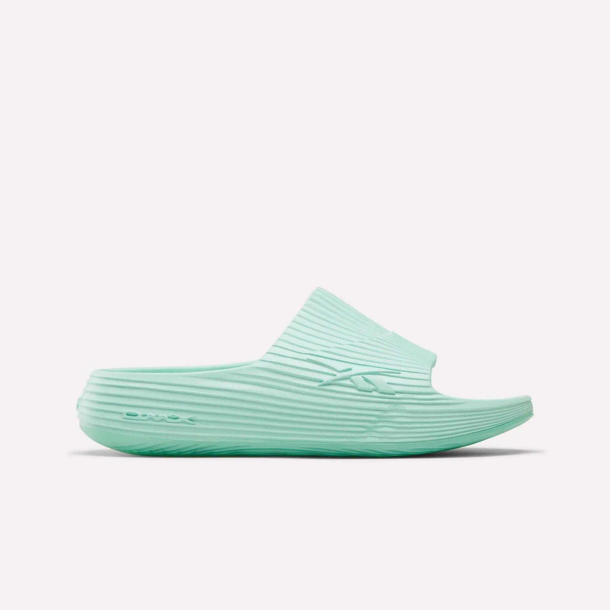 The Reebok DMX Recovery Walking Slides in light teal feature a ribbed texture, open toe, and thick sole with moving air cushioning, displayed in side profile on a white background.