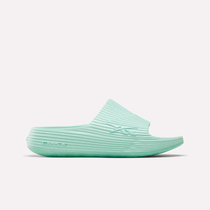 The Reebok DMX Recovery Walking Slides in light teal feature a ribbed texture, open toe, and thick sole with moving air cushioning, displayed in side profile on a white background.