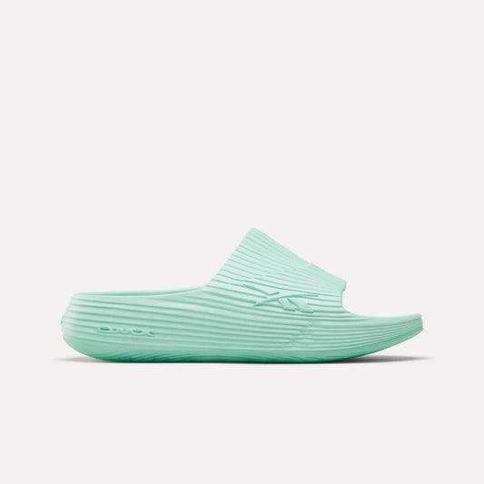 The Reebok DMX Recovery Walking Slides in light teal feature a ribbed texture, open toe, and thick sole with moving air cushioning, displayed in side profile on a white background.