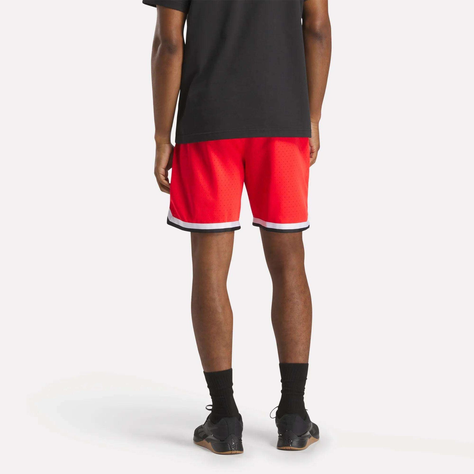 A person, seen from behind against a white background, wears a black t-shirt and Reebok Men's Basketball Transition Shorts 7" in red with white trim, paired with black socks and sneakers.