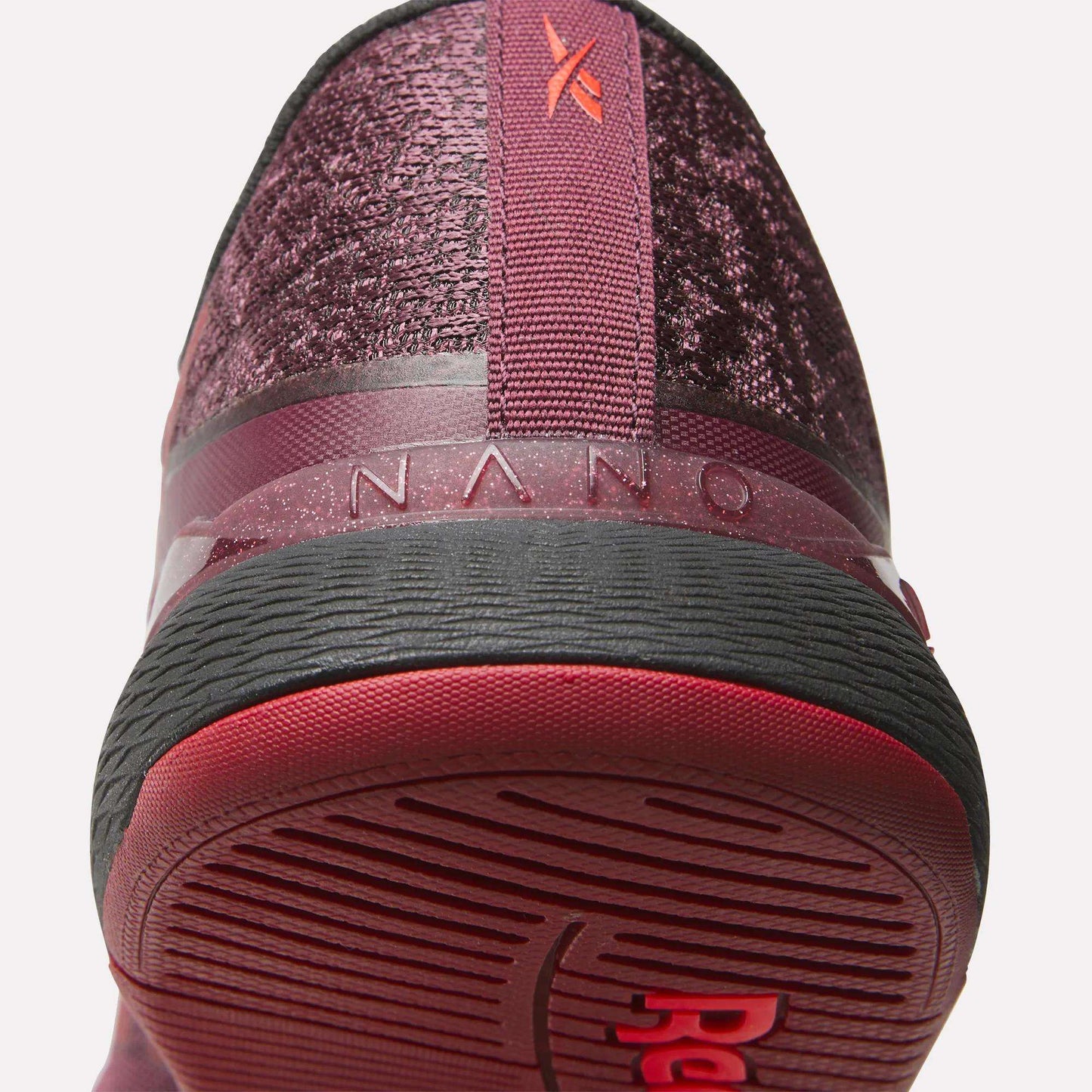 Close-up of the heel and sole of Reebok Women's Nano X5 Training Shoes in burgundy, featuring textured fabric, a red pull tab, "NANO" on the back, and a black and red rubber outsole for comfortable support.