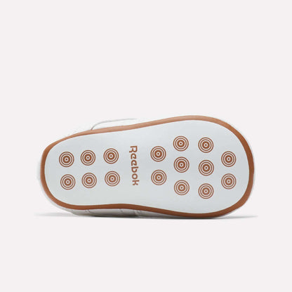 The image shows the sole of a white Kids' Club C Crib Shoe by Reebok for toddlers, featuring brown circular grip patterns and a brown Reebok logo at the center.