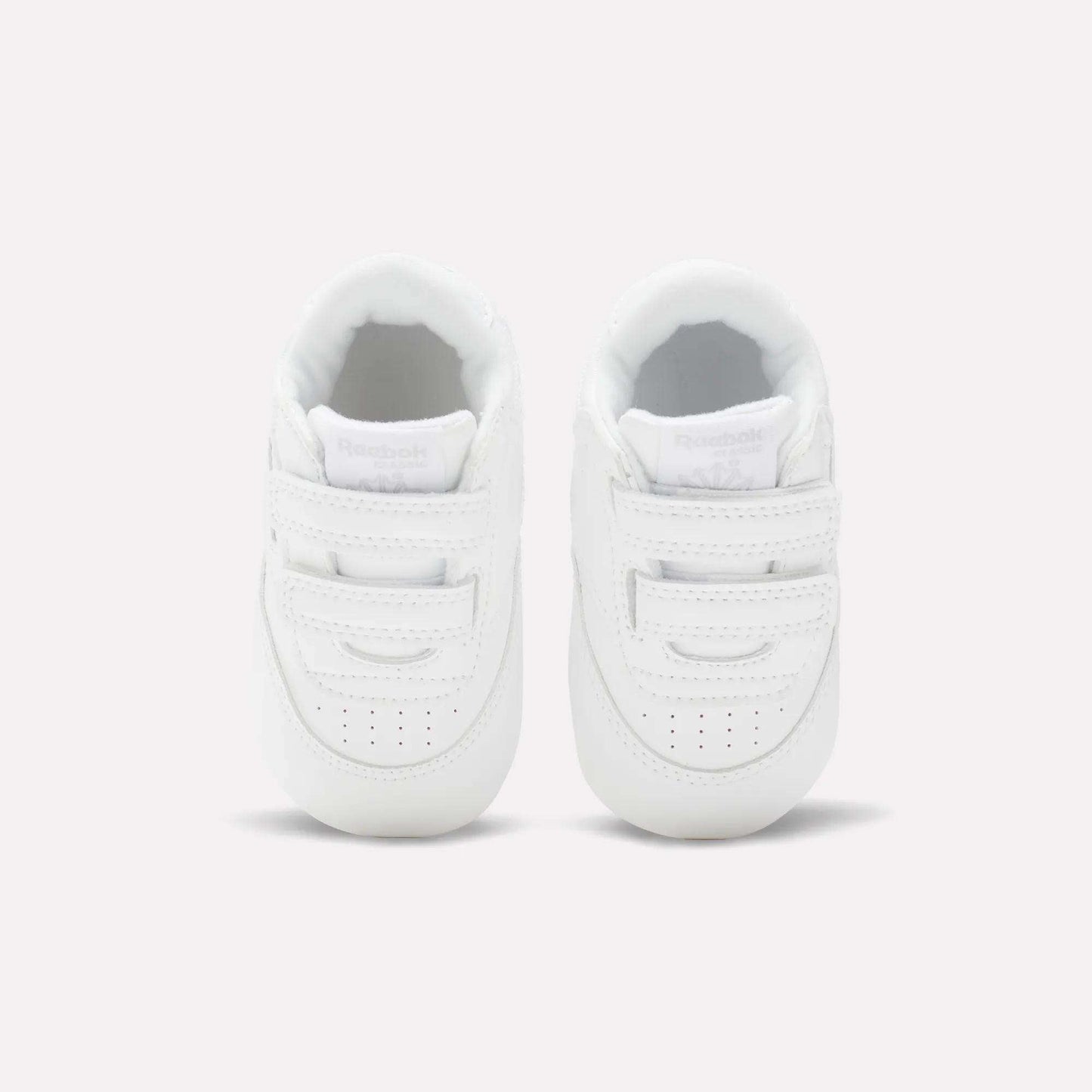 The Reebok Kids' Club C Crib Shoes for babies and toddlers are small, white shoes with hook-and-loop closures and a soft leather upper, shown from above on a plain light background.