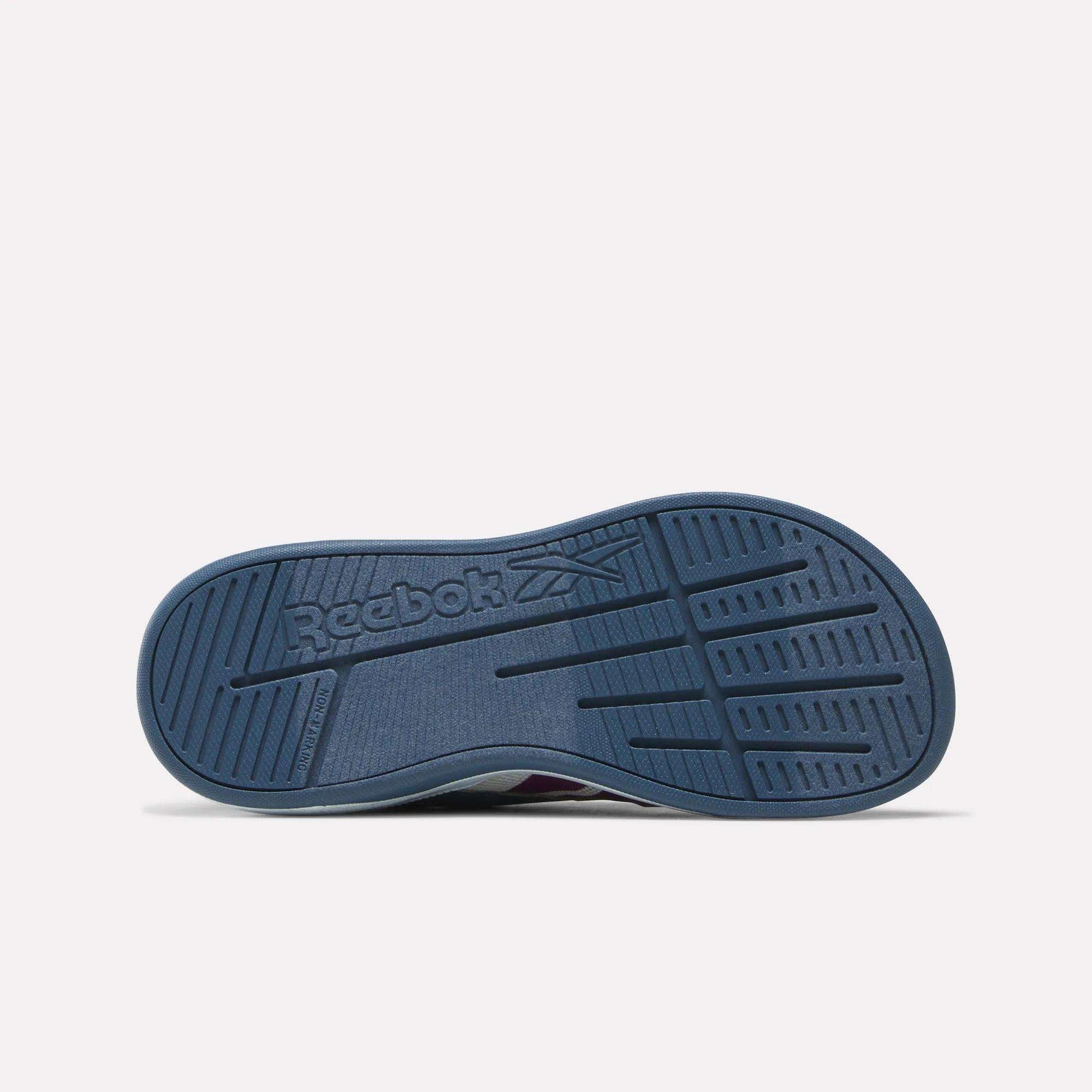 The image displays the blue rubber sole of the Reebok Kids' Nano Play Slip-on, viewed from below. The sole features multiple grooves along with the embossed Reebok logo and its signature vector symbol at the center.