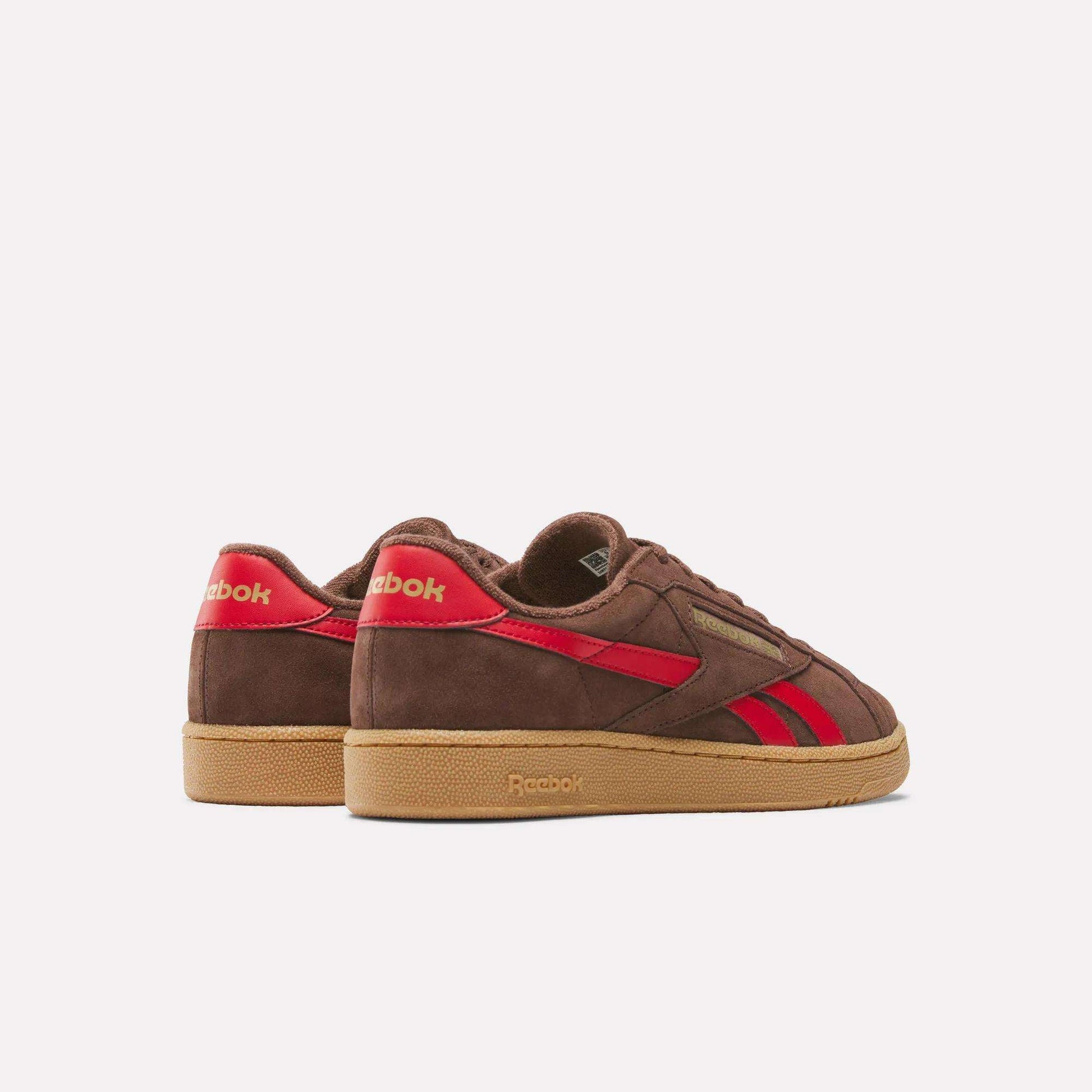 The Reebok Club C Grounds UK Shoes in brown feature red accents, a suede upper, and a gum sole, shown from a rear-side angle against a plain white background.