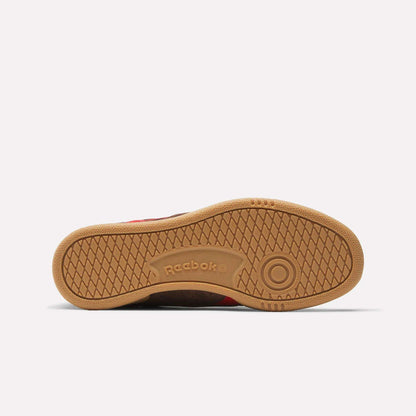 The image shows the sole of Reebok Club C Grounds UK Shoes, featuring a gum-colored rubber outsole with a circular toe pattern and herringbone grip, plus “Reebok” embossed near the arch to highlight the brand’s classic heritage.
