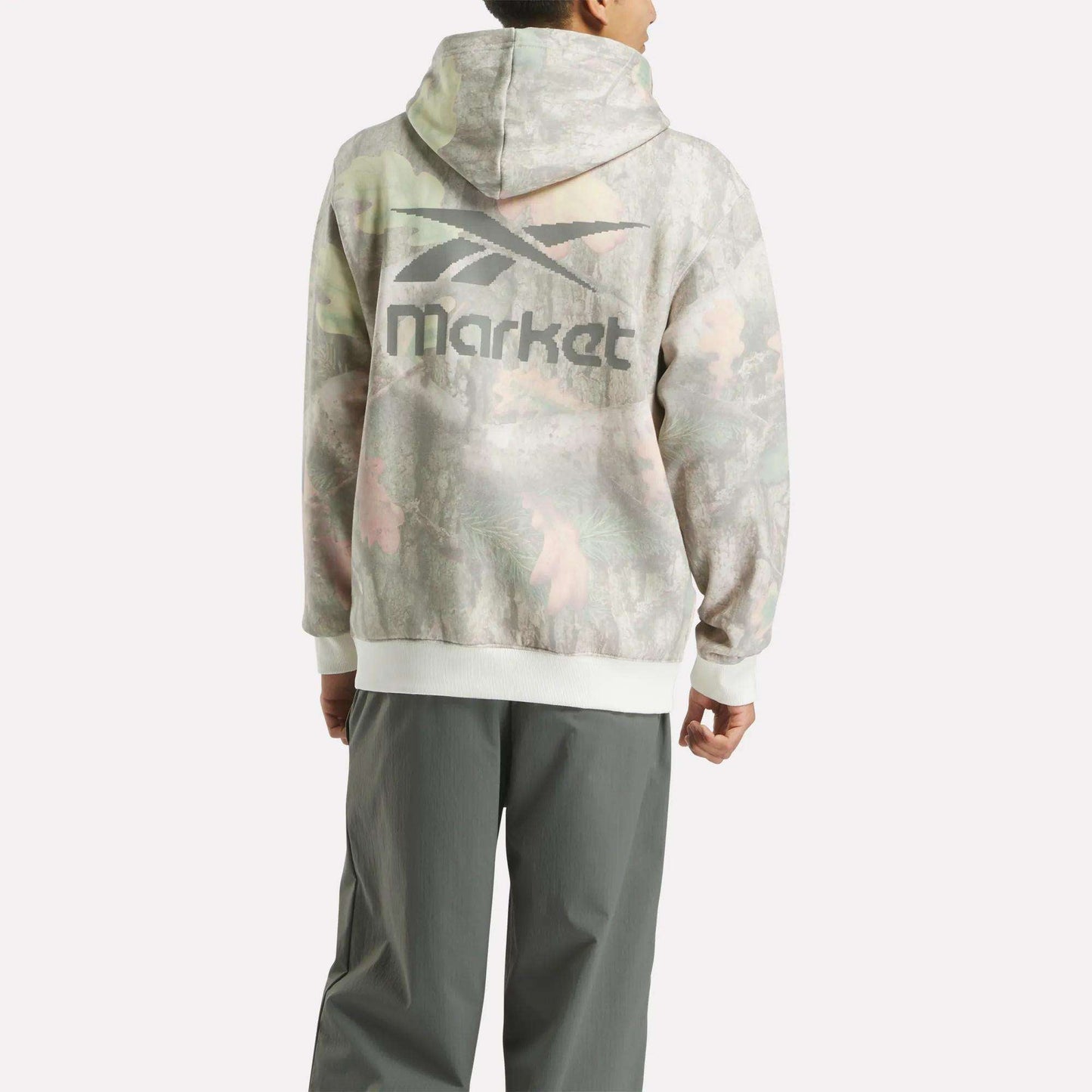 A person faces away, wearing a streetwear-inspired Reebok x MARKET Hoodie with a faded camo print and stylized logo on the back, paired with loose gray pants.