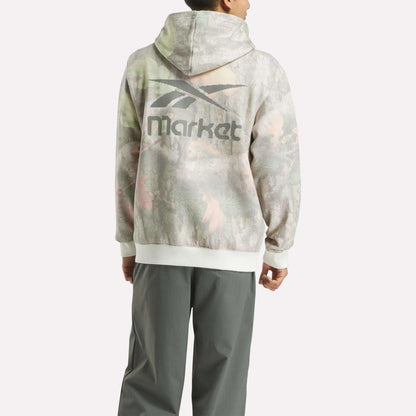 A person faces away, wearing a streetwear-inspired Reebok x MARKET Hoodie with a faded camo print and stylized logo on the back, paired with loose gray pants.