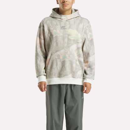 A person wearing a Reebok x MARKET Hoodie with a light camo print and hood, paired with gray pants, stands facing forward against a plain white background.