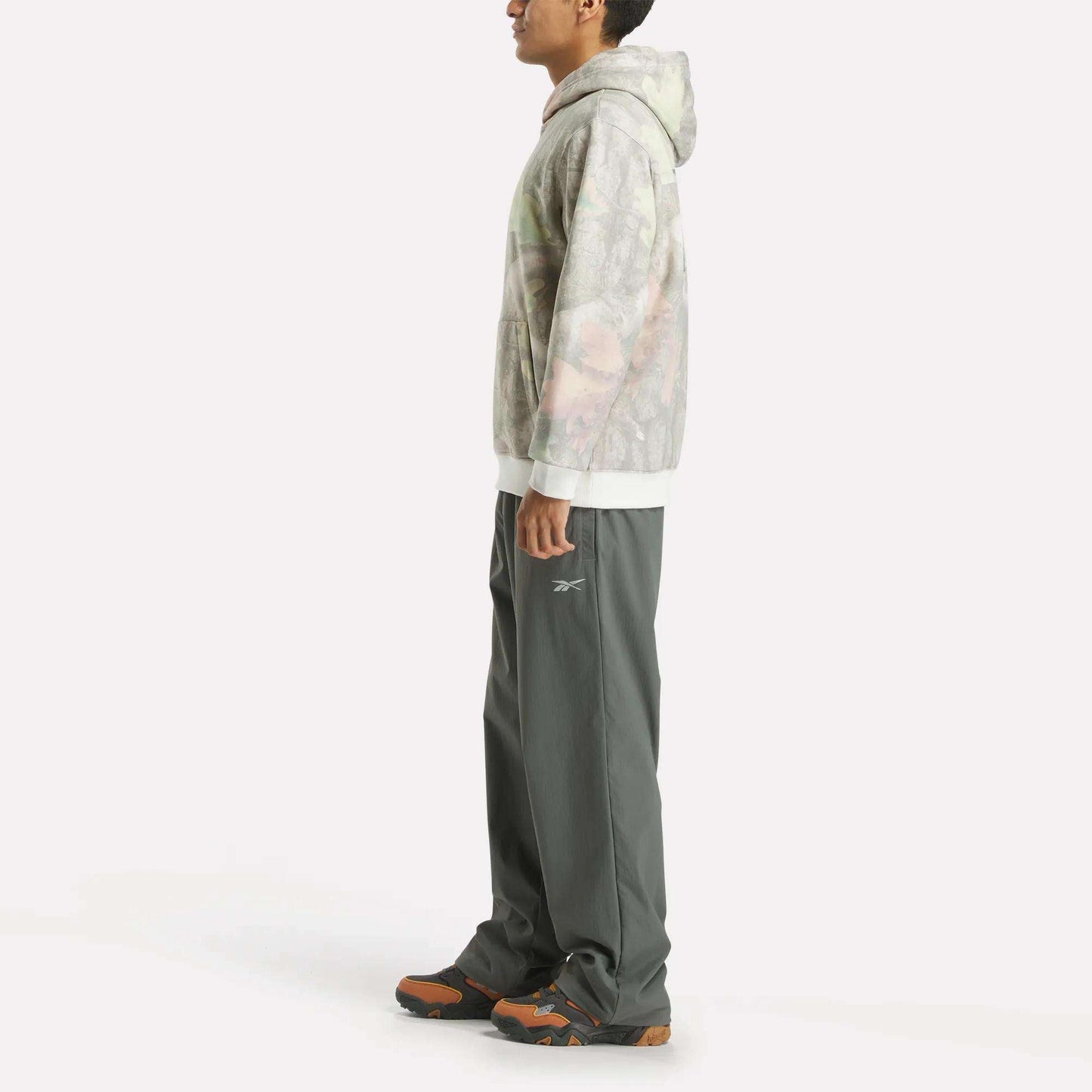 A person in profile wears the Reebok x MARKET Hoodie, loose gray pants with a small Reebok logo, and black-orange athletic shoes, styled against a plain white background for a streetwear-inspired look.
