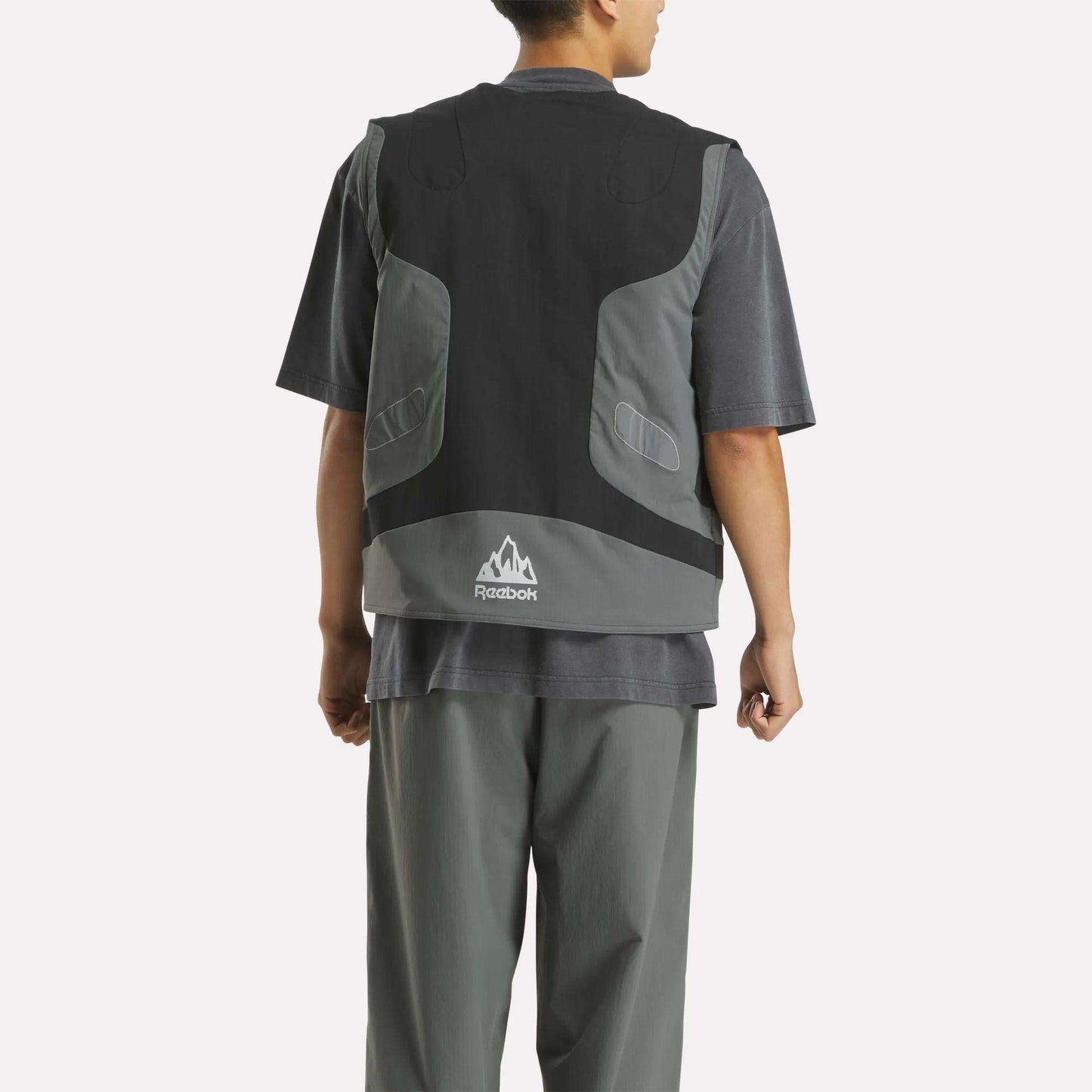 A person stands facing away, wearing the Reebok x MARKET Reversible Vest in black and gray over a dark tee and gray pants. The vest’s back features a small white mountain logo and Reebok branding, adding streetwear style.