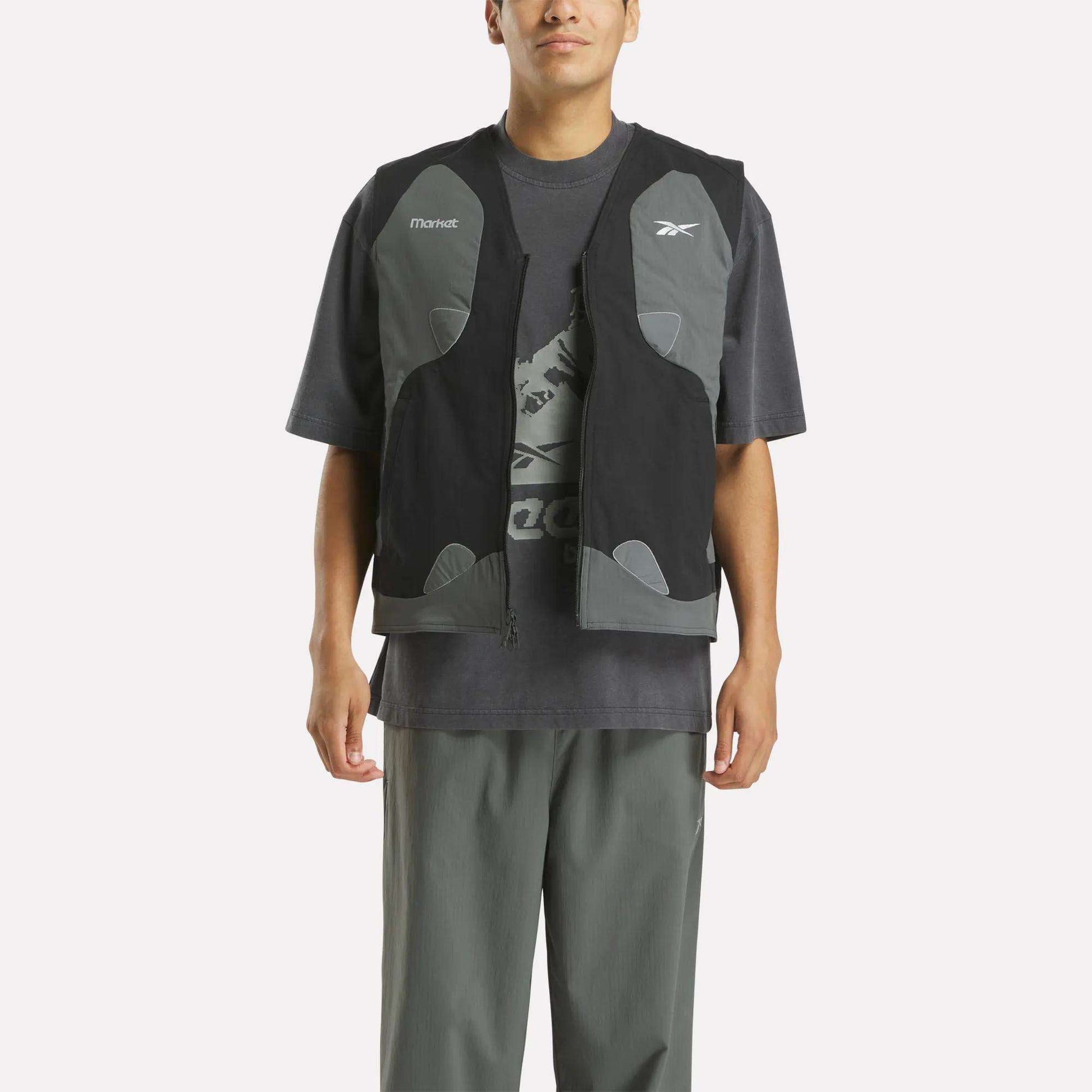 A person stands facing forward against a white background, wearing a loose dark gray Reebok T-shirt with a graphic logo, gray pants, and the Reebok x MARKET Reversible Vest in black and gray—capturing modern streetwear style with hands by their sides.