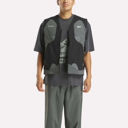 A person stands facing forward against a white background, wearing a loose dark gray Reebok T-shirt with a graphic logo, gray pants, and the Reebok x MARKET Reversible Vest in black and gray—capturing modern streetwear style with hands by their sides.
