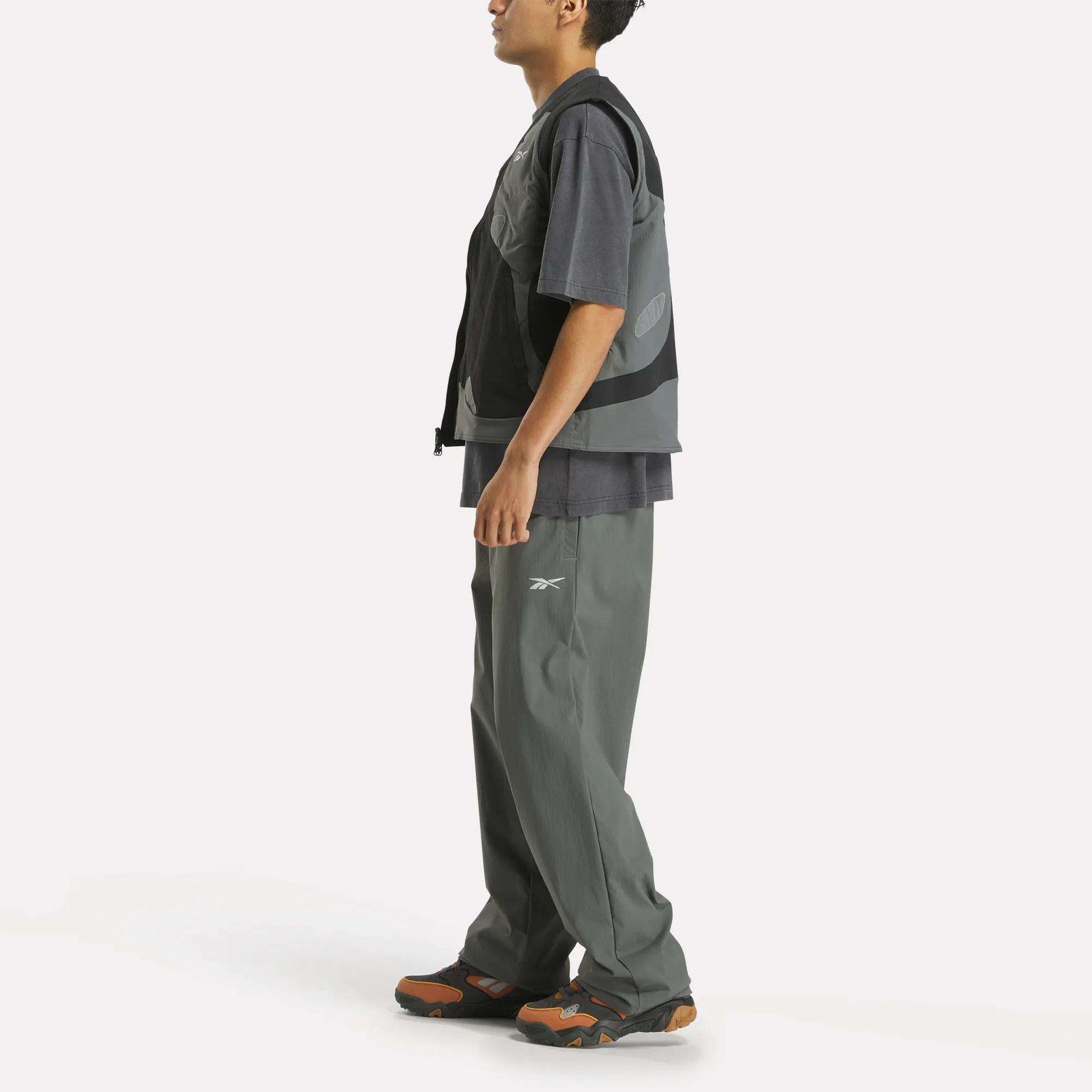 A person stands sideways against a white background, wearing a Reebok x MARKET Reversible Vest over a loose gray t-shirt, baggy gray Reebok pants, and sneakers with orange accents.