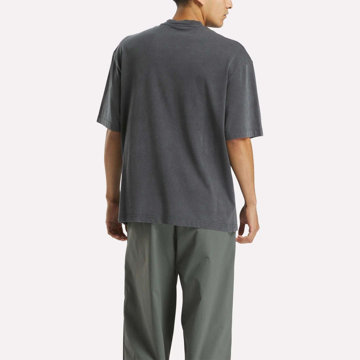 A person stands facing away in a loose, dark gray Reebok x MARKET Graphic T-Shirt and light gray pants against a plain white background, showcasing casual streetwear style.