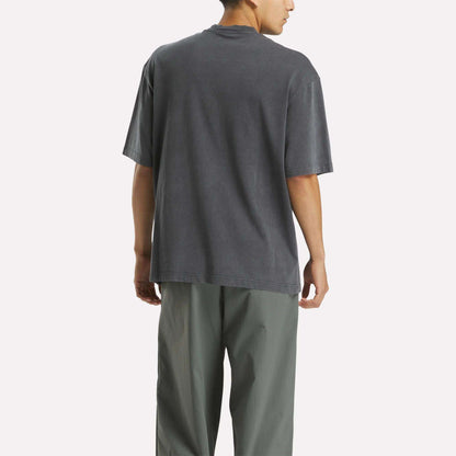 A person stands facing away in a loose, dark gray Reebok x MARKET Graphic T-Shirt and light gray pants against a plain white background, showcasing casual streetwear style.