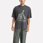 A person wears a loose-fitting Reebok x MARKET Graphic T-Shirt in dark gray with a bold logo and gray pants, standing against a plain white background for an effortless streetwear look.