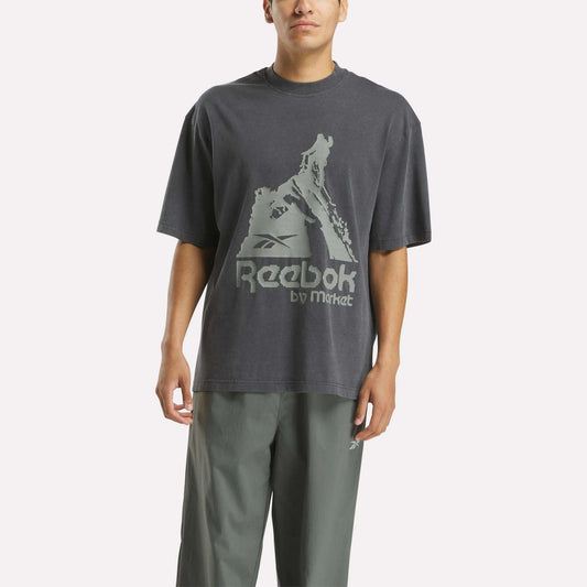 A person wears a loose-fitting Reebok x MARKET Graphic T-Shirt in dark gray with a bold logo and gray pants, standing against a plain white background for an effortless streetwear look.