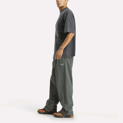 A person stands sideways in a relaxed streetwear look, wearing the Reebok x MARKET Graphic T-Shirt and dark green Reebok pants with black sneakers accented in orange, set against a plain white background.