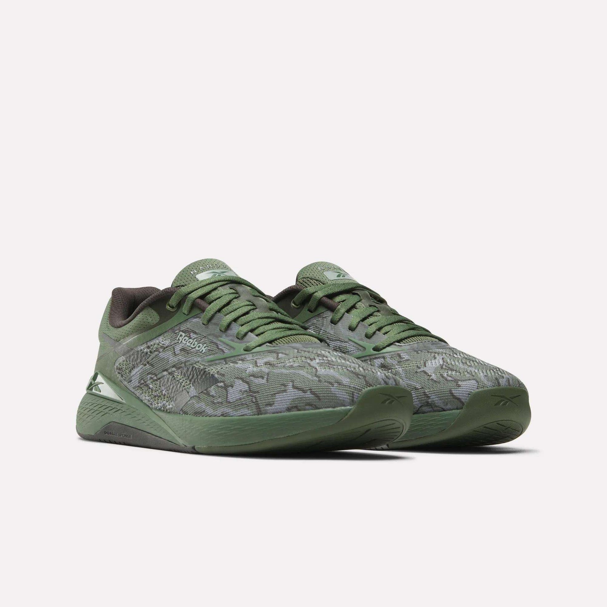 The Reebok Nano X5 Training Shoes for men feature a green camouflage pattern, dark green soles, and Reebok logos on the side and tongue. These iconic shoes are displayed against a plain white background.