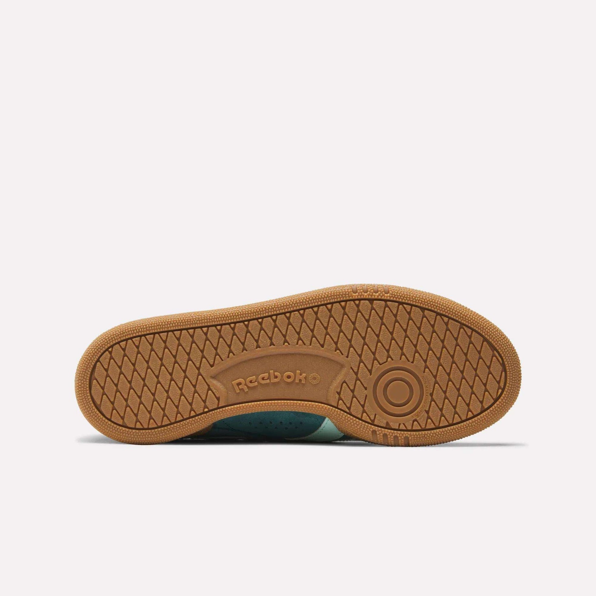 The image displays the bottom view of Reebok Women's Club C Grounds UK Shoes, showcasing a brown rubber sole with a circular grip pattern and the embossed Reebok logo in the center, emphasizing their iconic style.
