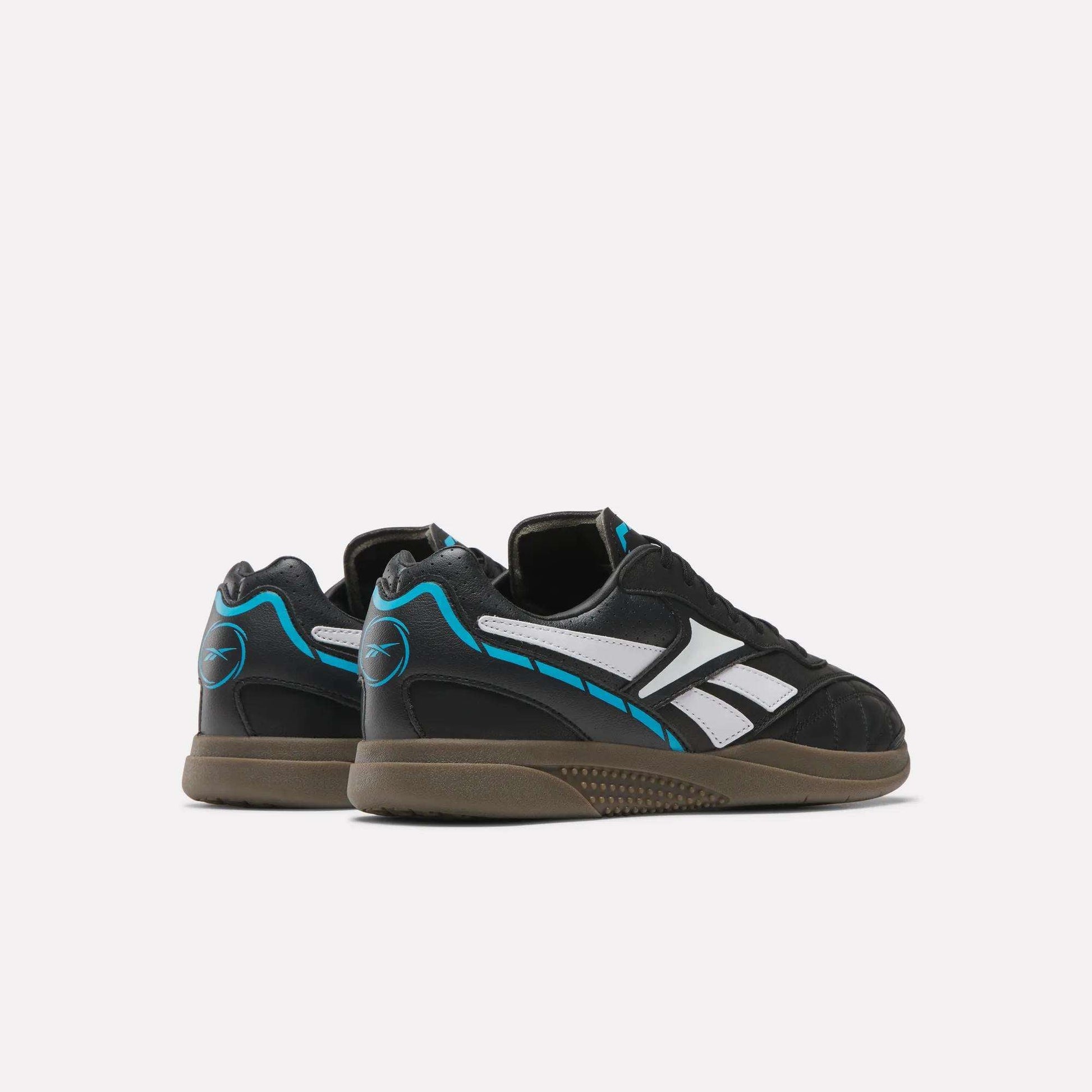 A pair of Reebok Hammer Street Shoes is shown against a plain white background, featuring black uppers with white stripes, blue accents, and brown soles. The soccer-inspired shoes are angled to display both the back and side views.