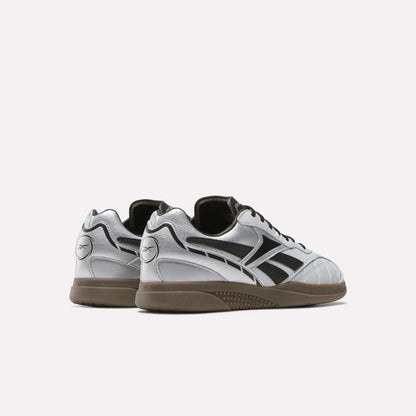 The Reebok Hammer Street Shoes in silver and black feature dark soles, black logo accents, full-grain leather panels, and detailed stitching. Pictured from the back and side against a white background, these sleek shoes offer a soccer-inspired look.