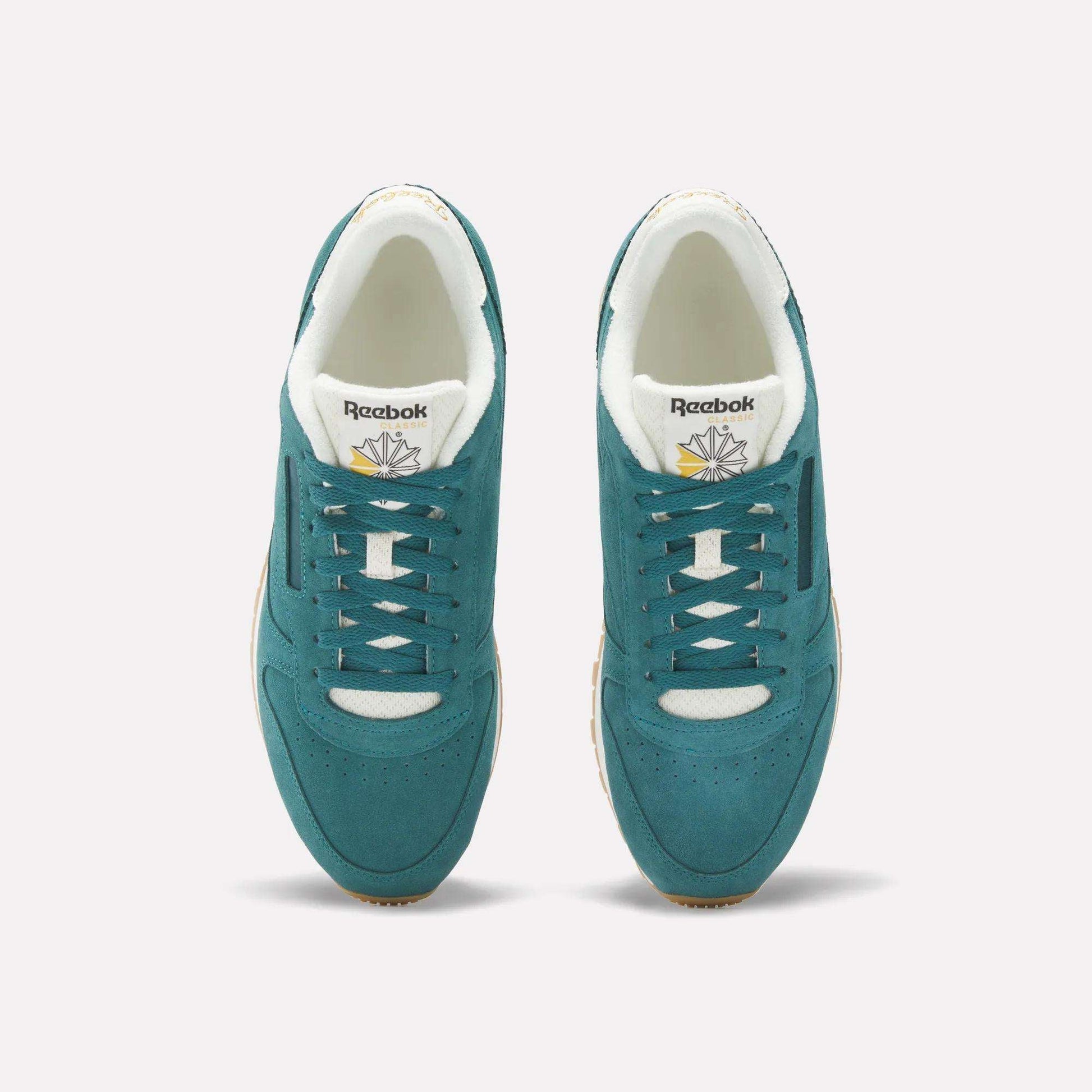 A pair of Classic Leather Shoes by Reebok in teal suede with white accents, shown from above. The image highlights the laces, gold and white branded tongue, beige outsoles, and a light background.