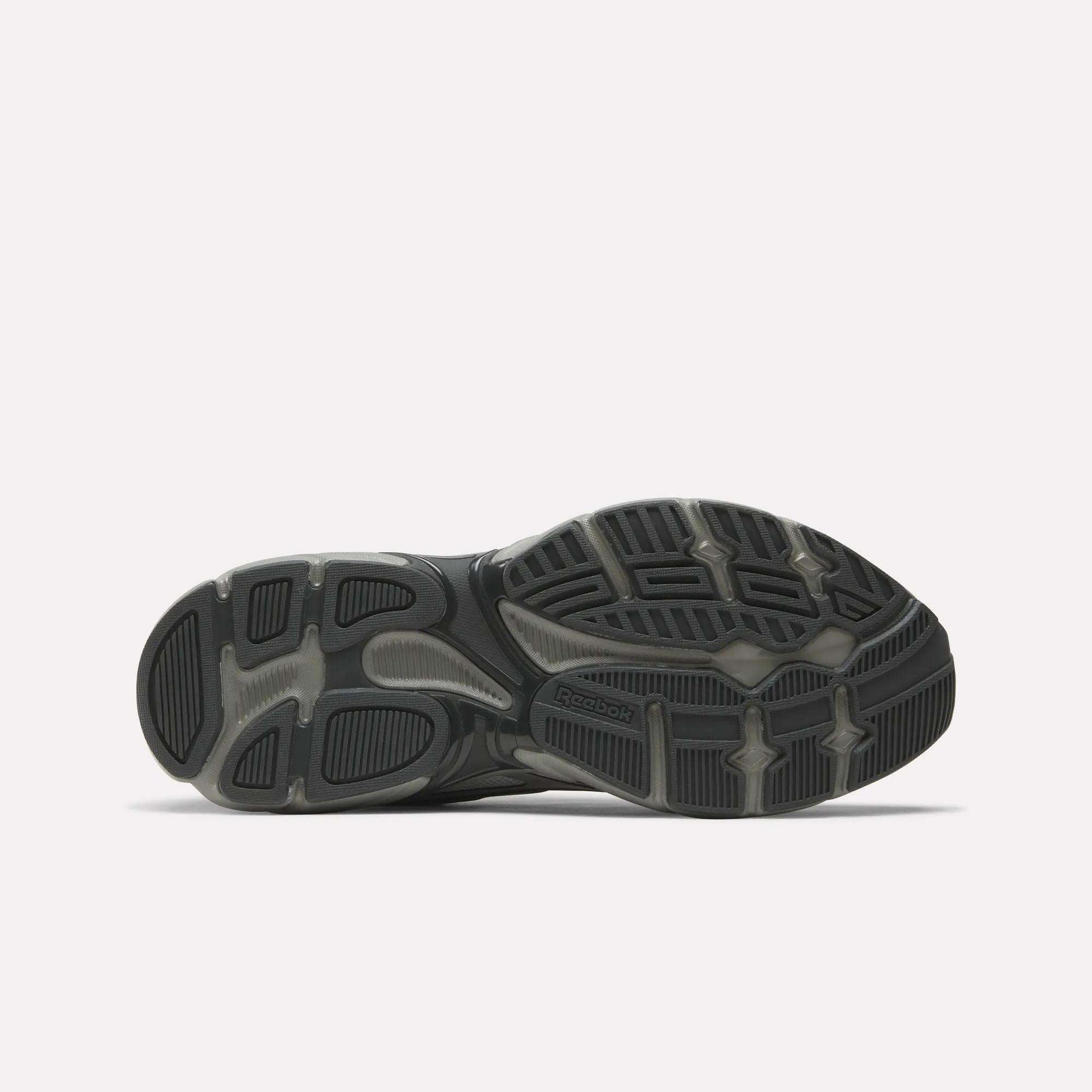 The image shows the sole of Reebok Premier Road Control Shoes, mainly black and gray, with textured rubber patterns and grooves for traction. The Reebok logo appears near the center, reflecting these classic retro running shoes.