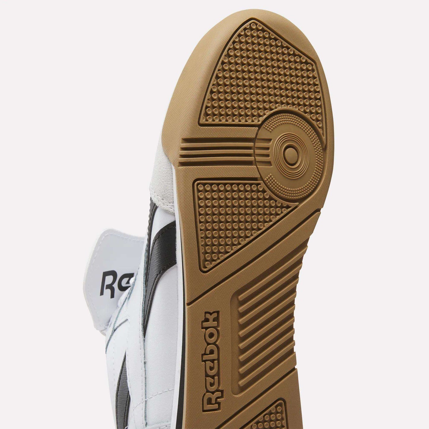 Close-up of the sole of Reebok Campio XT Kilty Shoes, featuring a brown rubber outsole with textured grip and embossed logo. The tennis-inspired design stands out with a white upper and black side stripe.