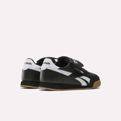 A pair of Reebok Campio XT Kilty Shoes in black with white stripes, white heel accents, gum soles, and velcro straps are displayed on a light background, angled to showcase the back and side tennis-inspired design.