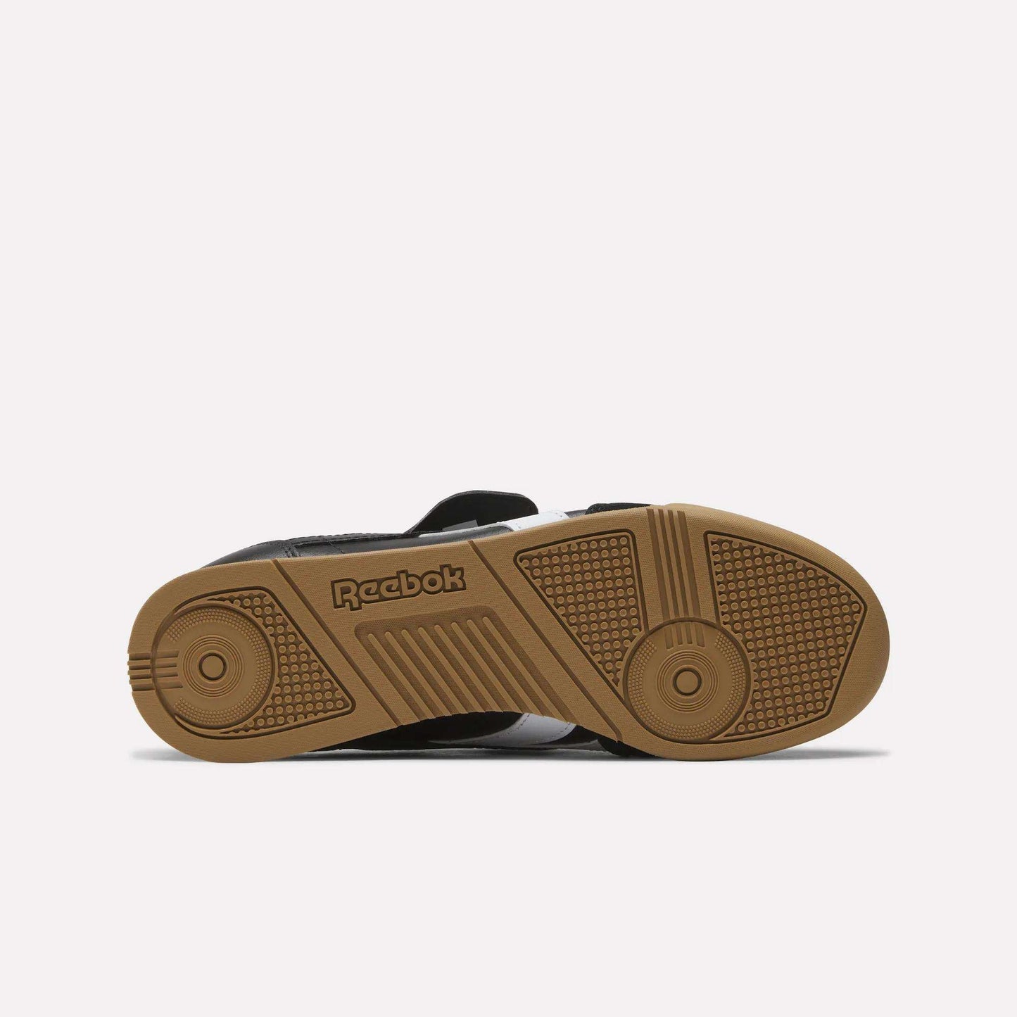 The image displays the sole of a black Reebok Campio XT Kilty Shoe with a gum-colored bottom, featuring tennis-inspired style, a textured grip pattern, and an embossed Reebok logo that blend function and fashion.