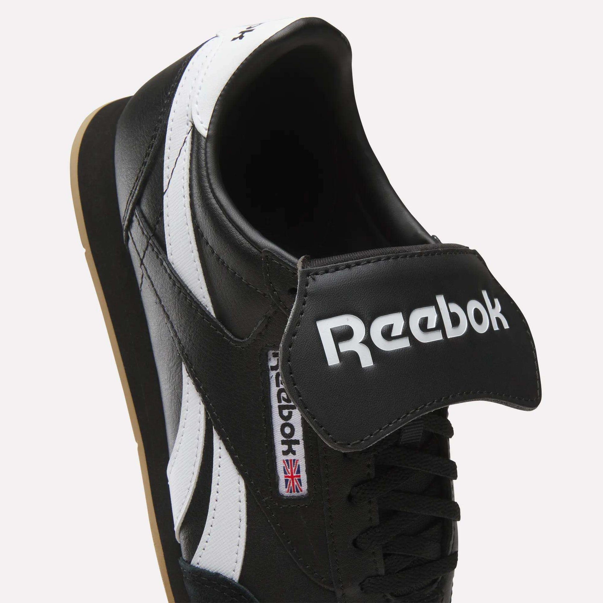 A close-up of the Reebok Campio XT Kilty Shoes in black leather with white stripes, featuring tennis-inspired style, a bold Reebok logo on a large tongue flap, and a small UK flag tag on the side.