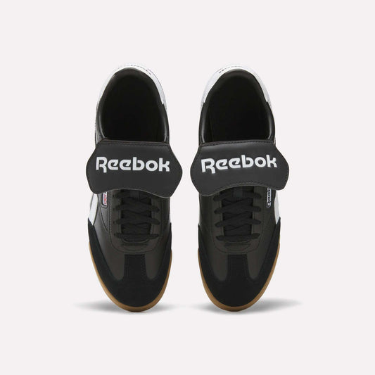 Top view of Reebok Campio XT Kilty Shoes in black with wide Velcro straps, white Reebok logo, black laces, and light brown soles. These tennis-inspired sneakers are shown on a plain white background.