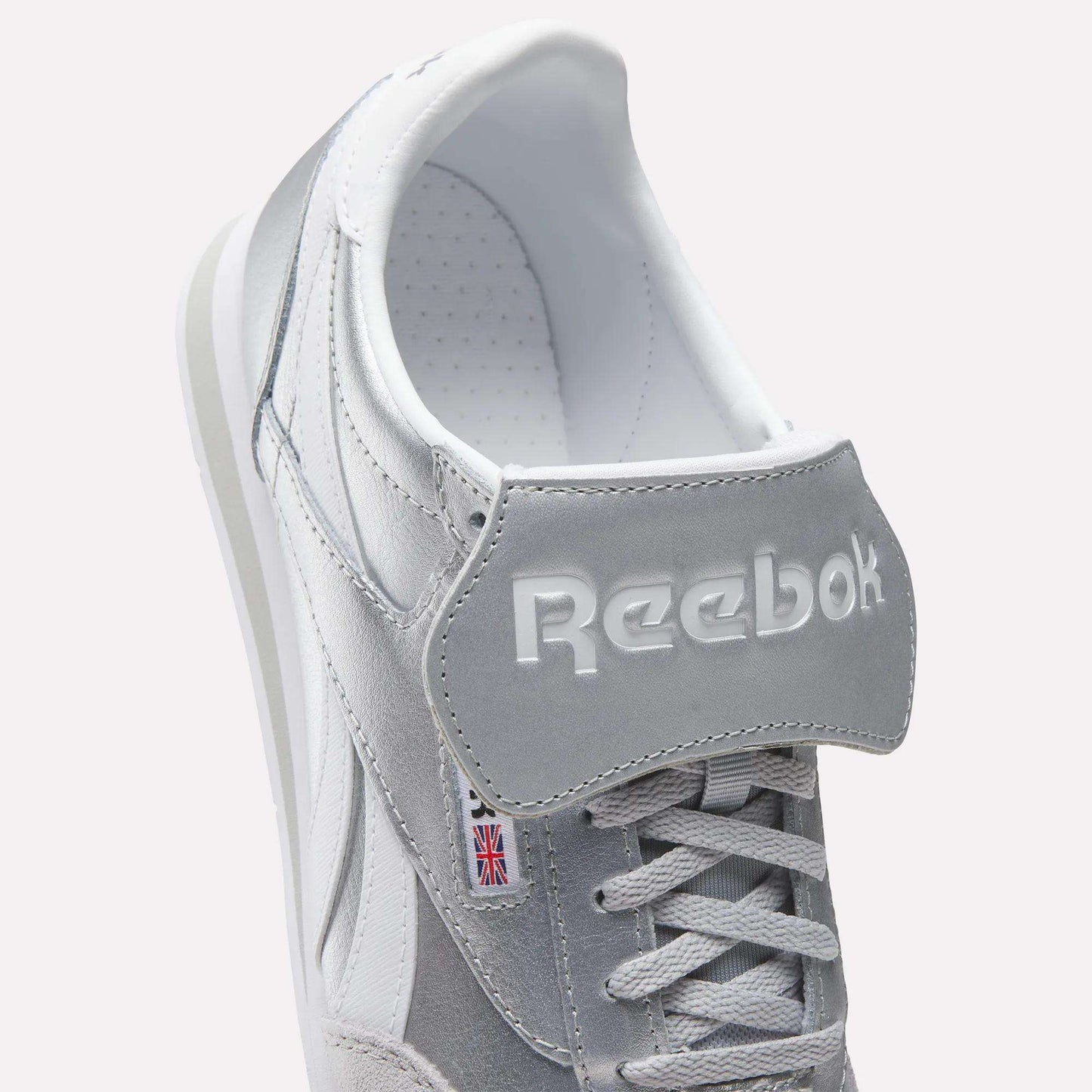 Close-up of Reebok Campio XT Kilty Shoes in gray and white with white laces, embossed Reebok logo on the tongue, and a small Union Jack label on the side, highlighting their classic tennis-inspired style.