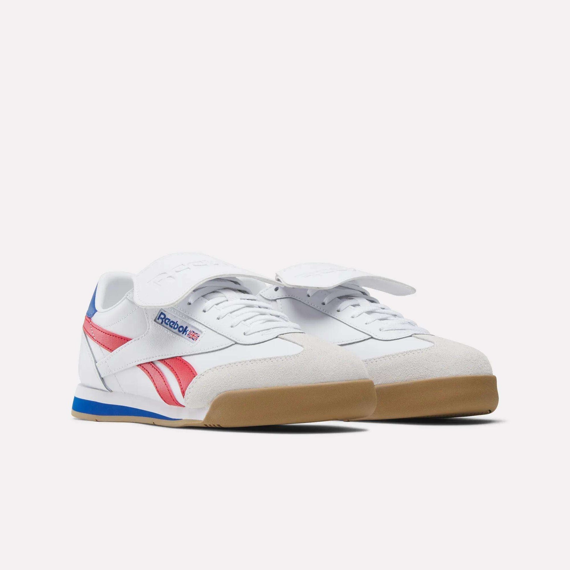 The Reebok Campio XT Kilty Shoes feature a white base with red and blue accents, beige suede toe detailing, gum soles, and a bold white strap over the laces, styled against a plain white background for tennis-inspired flair.