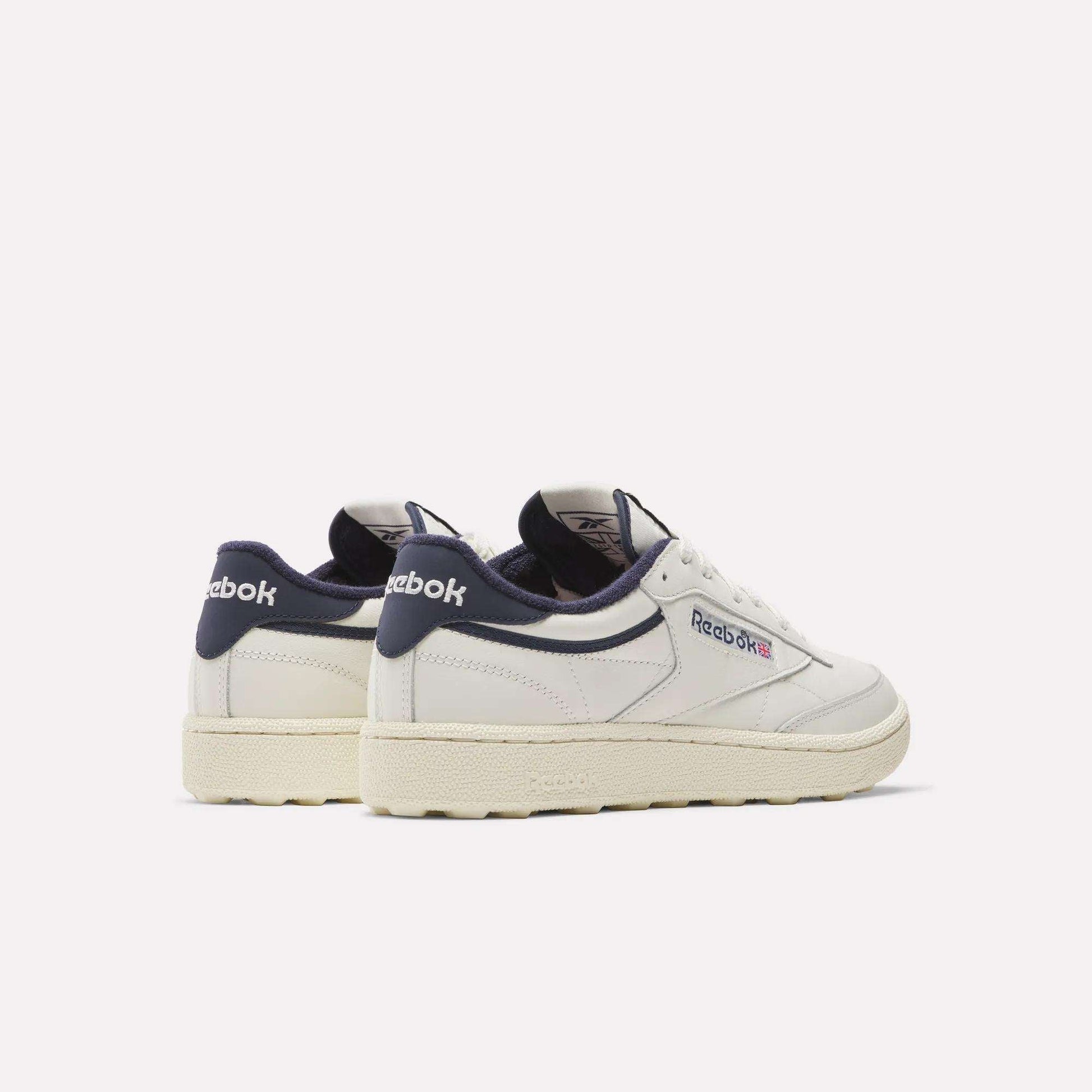 A pair of Reebok Men's Club C Golf Shoes in white with navy accents and off-white soles, featuring water-resistant leather uppers and a spikeless outsole, displayed on a plain light background, one showing the back and the other the side.