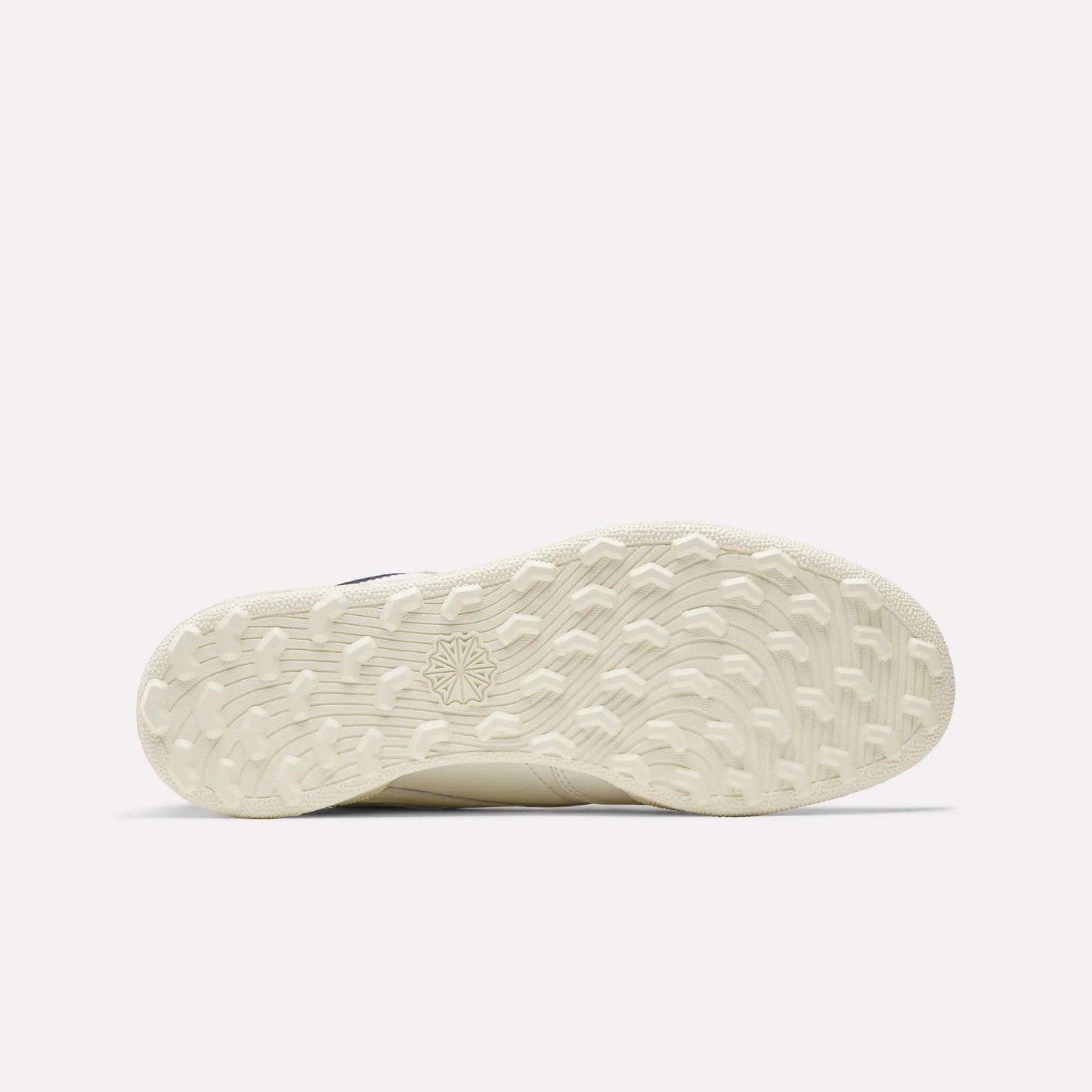 The image shows the off-white, textured sole of the Reebok Men's Club C Golf Shoes. It features a spikeless outsole with a patterned grip and starburst design near the center. The background is plain and light colored.