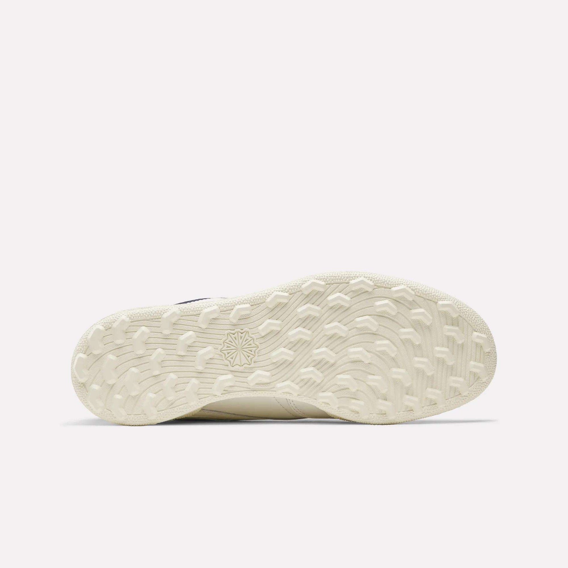 The image shows the off-white, textured sole of the Reebok Men's Club C Golf Shoes. It features a spikeless outsole with a patterned grip and starburst design near the center. The background is plain and light colored.