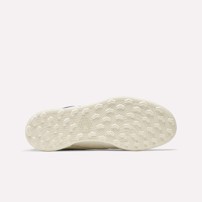 The image shows the off-white, textured sole of the Reebok Men's Club C Golf Shoes. It features a spikeless outsole with a patterned grip and starburst design near the center. The background is plain and light colored.